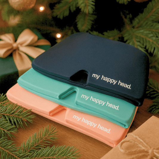 Tre My Happy Head huvudvärksmössor i svart, turkos och rosa ligger staplade som julklappsinspiration framför en gran.