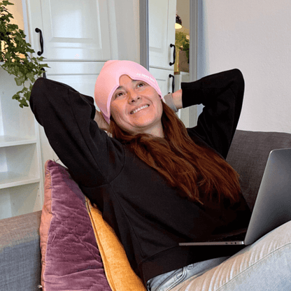 Leende person med laptop i knät som slappnar av hemma med rosa huvudvärksmössa från My Happy Head som ger kylande återhämtning och avslappning