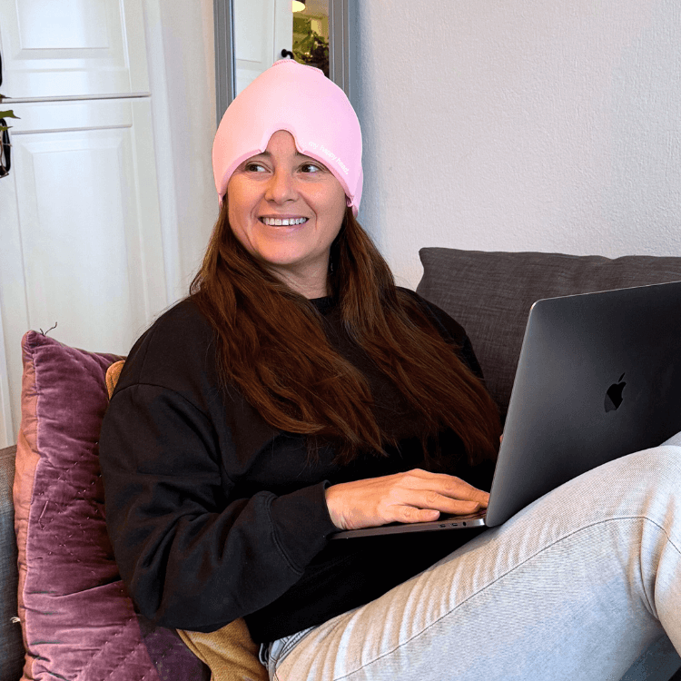Leende person som arbetar med laptop hemifrån med rosa huvudvärksmössa från My Happy Head för att minska spänningar och främja avslappning.